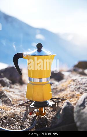 Caffè giallo moka pot con vapore al campeggio neve montagna. Picnic mattutino, persona che cucinano una bevanda calda per colazione nella natura. Scatto verticale Foto Stock