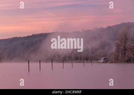 Mattina presto d'inverno durante un'alba vibrante piena di colori e con un velo di nebbia sul lago. I pali da pesca creano piacevoli riflessi Foto Stock