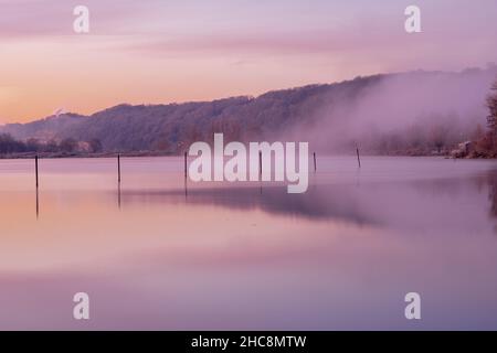 Mattina presto d'inverno durante un'alba vibrante piena di colori e con un velo di nebbia sul lago. I pali da pesca creano piacevoli riflessi Foto Stock