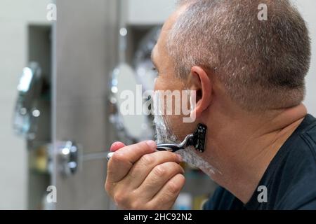 Uomo maturo dai capelli grigi che restyling la sua barba in casa utilizzando il rasoio Foto Stock