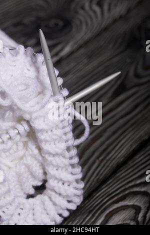 Lavorazione a maglia con aghi. Una matassa di filo bianco e un frammento di prodotto lavorato a maglia. Su assi di pino nero. Primo piano. Foto Stock