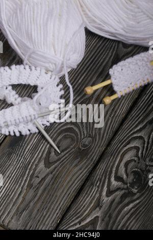 Lavorazione a maglia con aghi. Una matassa di filo bianco e un frammento di prodotto lavorato a maglia. Su assi di pino nero. Primo piano. Foto Stock