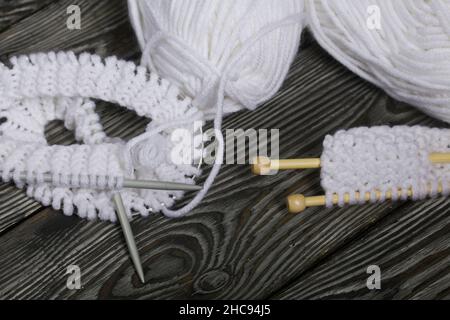 Lavorazione a maglia con aghi. Una matassa di filo bianco e un frammento di prodotto lavorato a maglia. Su assi di pino nero. Primo piano. Foto Stock