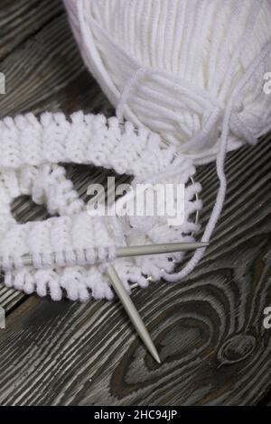 Lavorazione a maglia con aghi. Una matassa di filo bianco e un frammento di prodotto lavorato a maglia. Su assi di pino nero. Primo piano. Foto Stock