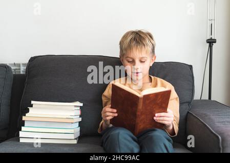 Adorabile ragazzo smart con capelli biondi in abiti casual lettura interessante finzione mentre seduto sul comodo divano vicino stack di vari libri Foto Stock