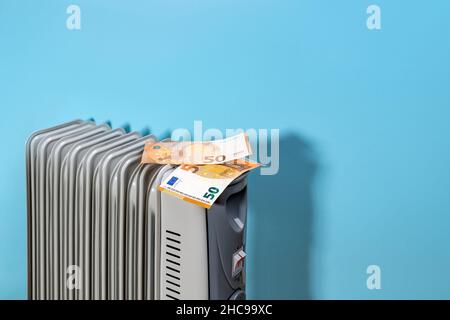 Cinquanta banconote in euro su un riscaldatore elettrico portatile riempito di olio vicino alla parete blu. Concetti di riscaldamento domestico, elettricità costosa e costo di elettricità. Foto Stock