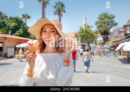 Una donna allegra con un sorriso bianco-neve mangia un delizioso gelato e riposa in una località turistica vicino al mare. Foto Stock