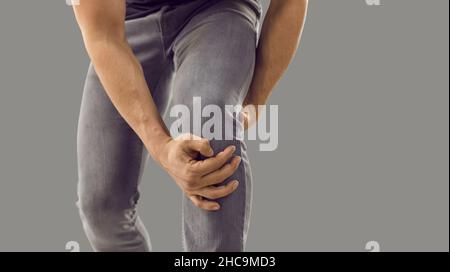 Primo piano di un uomo sconosciuto con trauma alle gambe che tiene il ginocchio ferito su sfondo grigio. Foto Stock