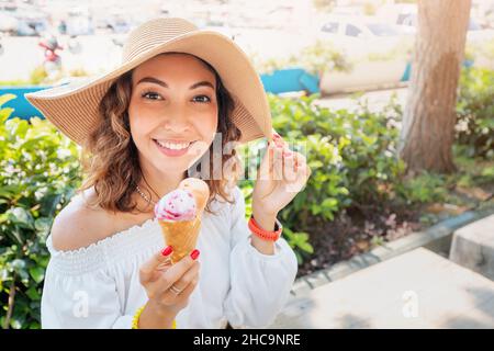 Una donna allegra con un sorriso bianco-neve mangia un delizioso gelato e riposa in una località turistica vicino al mare. Foto Stock