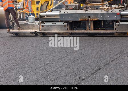 Un'asfaltatrice industriale stesa uno strato di nuovo asfalto caldo sulla carreggiata. Gli operatori controllano l'uniformità e l'altezza della pavimentazione. Spazio di copia. Foto Stock