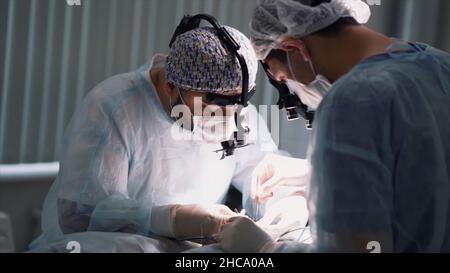 Team medico che esegue l'operazione. Primo piano dei volti dei chirurghi al lavoro indossando le feritoie binoculari in sala operatoria durante la procedura. Foto Stock