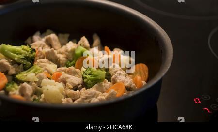 Cottura del pollo con le verdure in una padella nera in piedi su un piano di cottura sensibile al tatto. Preparazione di una cena sana, frittura di carote, broccoli e cauli Foto Stock