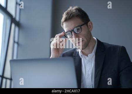 Concentrato giovane uomo d'affari tedesco in abito formale e occhiali guardando lo schermo del laptop Foto Stock