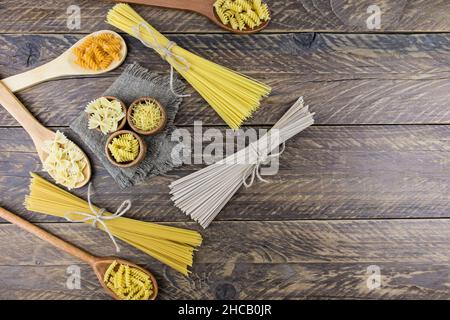 Vista dall'alto di vari tipi di pasta e spaghetti legati in un mazzo, in cucchiai e ciotole di legno su sfondo ligneo, copia dello spazio Foto Stock