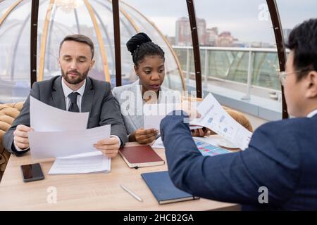 Due giovani colleghi seri che guardano attraverso i documenti in occasione di un incontro di lavoro con un partner commerciale straniero in caffetteria Foto Stock