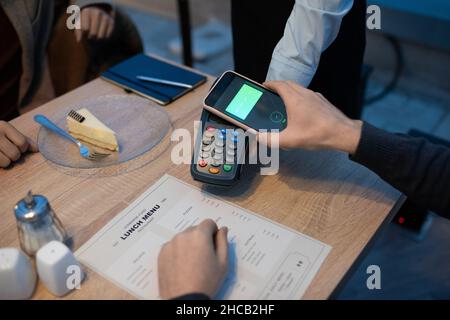 Mano di uomo d'affari che tiene smartphone sul terminale di pagamento mentre paga per il cibo ordinato nel ristorante Foto Stock