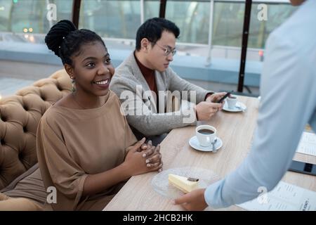Felice giovane donna africana in elegante casualwear guardando la cameriera mettere piatto con cheesecake sul tavolo in caffè Foto Stock