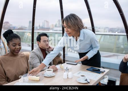 Giovane cameriera che si piega sul tavolo e che mette piatto con gustoso cheesecake di fronte alla donna africana Foto Stock