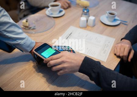 Giovane uomo che tiene smartphone sopra il terminale di pagamento tenuto dalla mano della cameriera mentre si siede da un tavolo di legno in un caffè Foto Stock