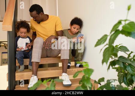 Giovane uomo di etnia africana che parla con uno dei suoi simpatici figli con smartphone mentre si siede su gradini di scala Foto Stock