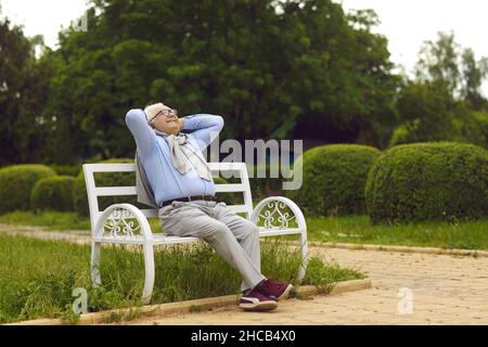 Uomo anziano sorridente felice respirando aria fresca seduto su una panca in un parco estivo verde Foto Stock