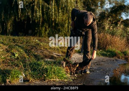 Il pescatore con asta rotante dà al gatto un persico di pesce sullo sfondo della natura. Uomo pescatore con pesca che gira o casting canna vicino al fiume. Pescatore Foto Stock