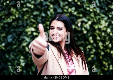Donna d'affari sorridente che mostra i pollici di fronte alle piante d'edera Foto Stock