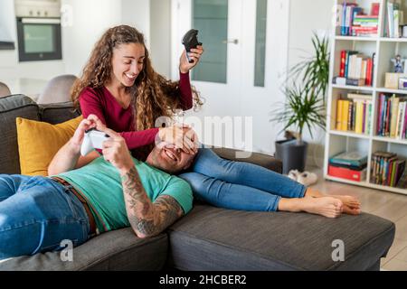 Donna sorridente con controller di gioco che copre gli occhi dell'uomo sul divano a casa Foto Stock