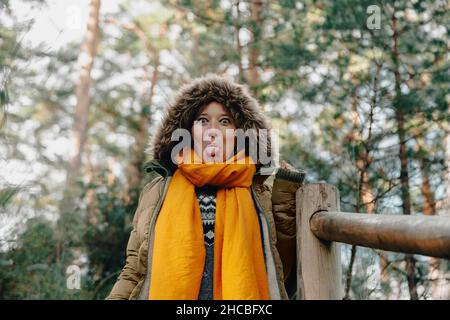 Donna in pelliccia cappuccio che sporge lingua da ringhiera a foresta Foto Stock