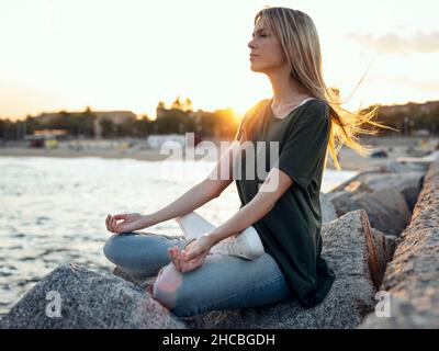 Donna bionda meditante sulla roccia a Bogatell Beach, Barcellona, Catalogna, Spagna Foto Stock