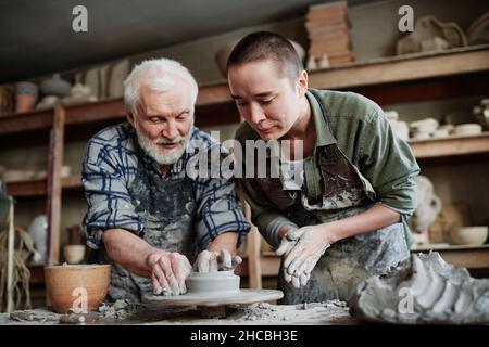 Uomo anziano che mostra alla donna come fare la forma di vaso da argilla su ruota in ceramica in studio Foto Stock