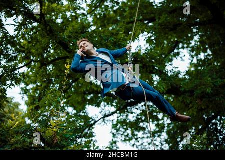 Un uomo d'affari sorridente appeso alla corda che parla al telefono cellulare al parco Foto Stock