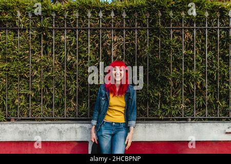 Donna felice con capelli rossi in piedi di fronte alla recinzione Foto Stock