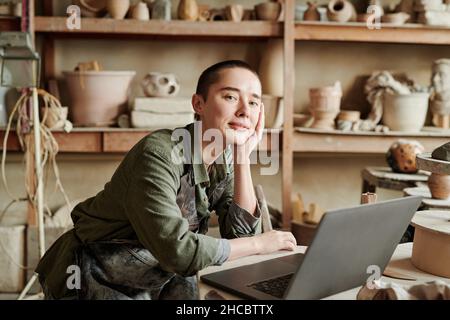 Ritratto di giovane artigiano che guarda la fotocamera mentre lavora su un computer portatile sul posto di lavoro in studio Foto Stock