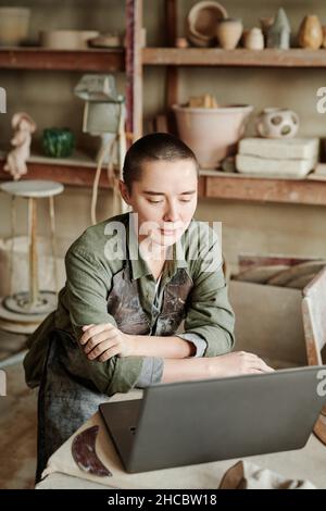 Giovane scultore serio che usa il laptop per sviluppare un nuovo progetto seduto al tavolo nel laboratorio di ceramica Foto Stock