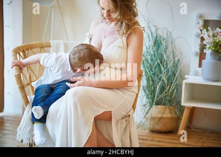 Madre incinta e figlio abbracciano e trascorrono il tempo insieme a casa. Un ragazzino bacia il pancino incinta di sua madre. Concetti di gravidanza, famiglia, parentodio Foto Stock