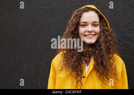 Primo piano orizzontale medio di donna caucasica che indossa un impermeabile giallo in piedi contro una parete grigio scuro guardando la fotocamera Foto Stock