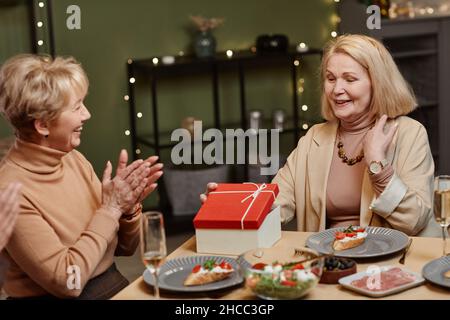 Allegra vecchia donna caucasica che apre la scatola presente mentre si siede al tavolo da pranzo e che festeggia il Natale con gli amici Foto Stock
