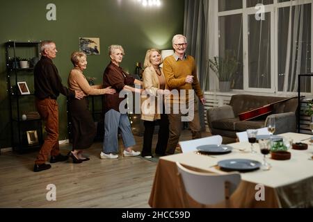 Due vecchi e tre donne che ballano e si divertono insieme in un soggiorno decorato con luci durante le riunioni di amici di festa Foto Stock
