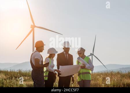 Team internazionale di business partner che si riuniscono tra turbine eoliche ed esaminano i progetti. Gruppo di professionisti che collaborano per la produzione di energia ecologica pulita. Foto Stock