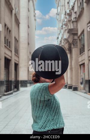 Donna nel mezzo di alcuni vecchi edifici prende il suo cappello nero che copre parte del suo volto. Spazio di copia Foto Stock