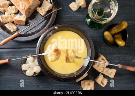 Fonduta di formaggio svizzero come pasto di festa di nuovo anno con pane su forchette lunghe, sottaceti e vino su un tavolo di legno scuro, alto angolo di vista dall'alto, selezionati foc Foto Stock