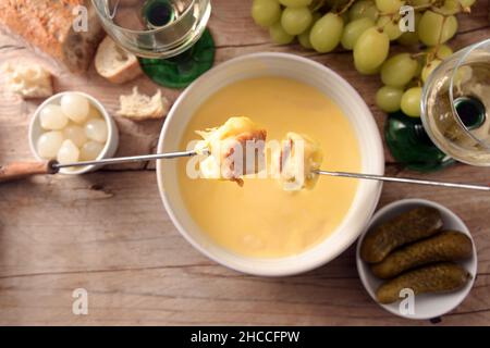 Fonduta di formaggio fuso con pane su forchette lunghe, sottaceti, uva e vino bianco su un rustico tavolo di legno, spazio copia, vista ad angolo alto dall'alto, selezionare Foto Stock