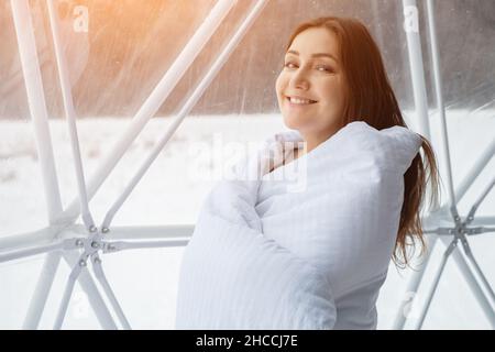 giovane donna in coperta sorridente si erge accanto alla finestra panoramica del campeggio, luce del sole Foto Stock