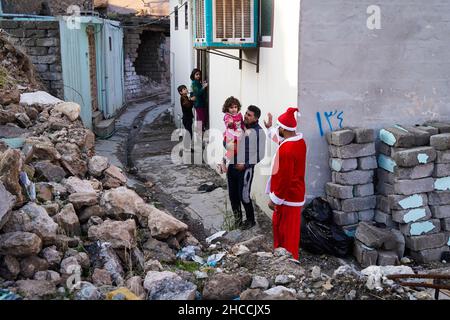 Mosul, Iraq. 24th Dic 2021. Un uomo vestito come Babbo Natale interagisce con i bambini dopo aver dato loro un albero vicino alle macerie nella città vecchia di Mosul.un'iniziativa lanciata da volontari iracheni dalla Mosul Eye Foundation per piantare migliaia di alberi nella città devastata dalla guerra di Mosul, Allo scopo di rendere più verde Mosul e combattere la desertificazione, distribuivano 100 alberi diversi alle case del centro storico di Mosul nell'ambito di un progetto volto a piantare 5.000 alberi in tutta la città settentrionale di Mosul, che soffre ancora per la distruzione e la devastazione lasciate dalla guerra contro Foto Stock