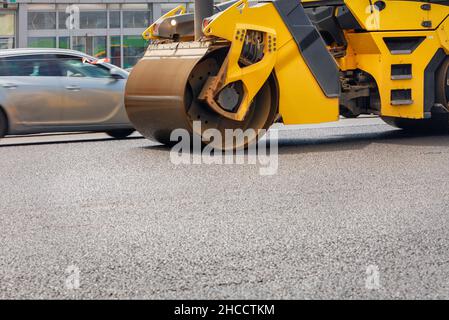 Un rullo giallo che costipa asfalto fresco su un tratto di strada recintato in una giornata estiva. Lieve sfocatura del movimento. Spazio di copia. Foto Stock