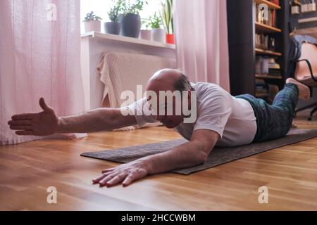 Uomo anziano che fa la variazione di Shalabhasana sul tappeto di esercitazione. Foto Stock