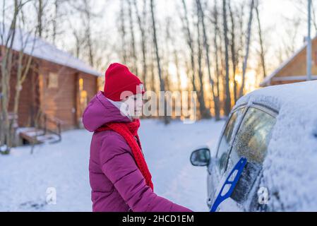 giovane donna in un cappello rosso che pulisce un'auto coperta di neve sullo sfondo di una strada invernale e il tramonto del sole. Foto Stock