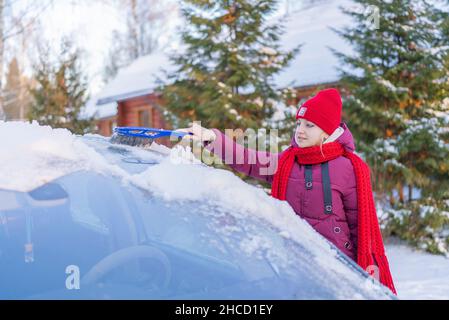 giovane donna in un cappello rosso che pulisce un'auto coperta di neve sullo sfondo di una strada invernale e il tramonto del sole. Foto Stock