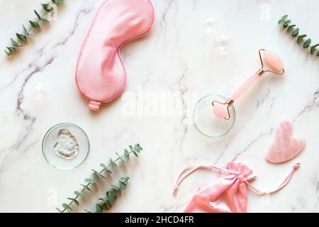 Moisturizer fatto a mano e rullo in pietra quarz rosa. Asciugamano di cotone. Foglie di eucalipto esotico e gocce d'acqua su sfondo di marmo bianco. Viso Foto Stock